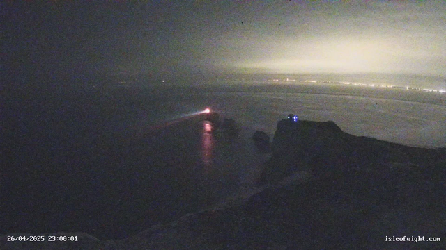 A dark, nighttime scene with a body of water reflecting faint lights. In the distance, a small boat or vessel is moving, emitting a bright red light that creates a trail on the water. The horizon shows a faint glow, possibly from distant city lights or stormy weather, and the sky appears overcast or foggy. The image has a timestamp in the lower left corner and a website in the lower right corner.