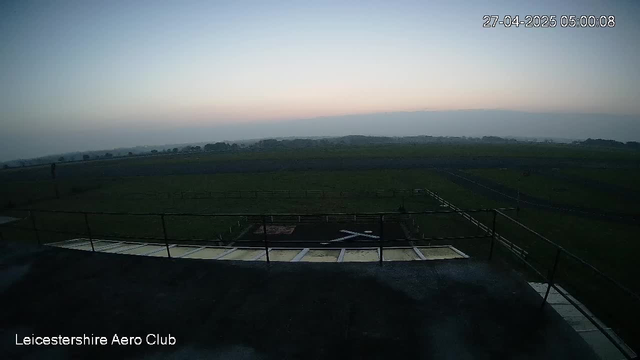 A view from a webcam showing an open field with green grass under a clear sky during dawn or dusk. In the foreground, there is a small helipad marked with an "H" and surrounded by a black railing. Text in the bottom left corner reads "Leicestershire Aero Club." The timestamp in the top right corner indicates the date as April 27, 2025, and the time as 05:00:08.