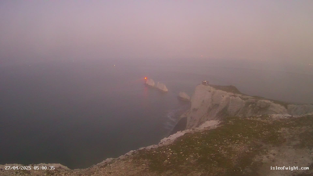 A view of white chalk cliffs overlooking the sea at dawn or dusk, with the coastline extending into the distance. Two small buildings are visible on the cliff tops, and a lighthouse with a red light is situated on a rock formation offshore. The sky appears hazy with a soft, muted color palette. The timestamp reads "27/04/2025 05:00:35" and the website "isleofwight.com" is displayed at the bottom right corner.