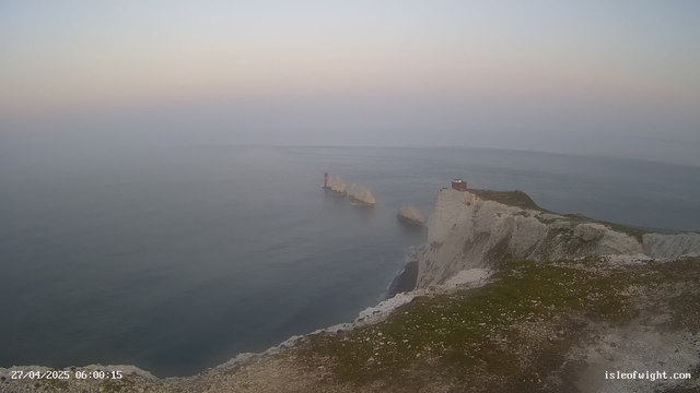 A view of white chalk cliffs with a rugged grassy top extending into the distance over a calm, greyish-blue ocean at dawn or dusk. There are large rock formations in the water near the base of the cliffs, and a small building and a walkway are visible atop the cliffs on the right side. The sky is clear with a gradient of pale colors, and the timestamp indicates 6:00 AM on April 27, 2025. The website is isleofwight.com.