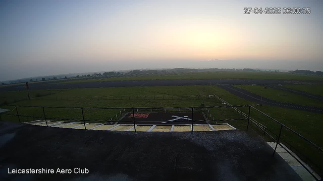 A wide view of an open grassy airfield at sunset, with a small helicopter landing pad in the foreground marked with a white "H" and a red border. The sky is clear with a faint glow from the setting sun, and there are distant low hills and open fields stretching toward the horizon. The timestamp in the upper right corner reads "27-04-2025 06:00:35," and the bottom left corner displays the text "Leicestershire Aero Club."
