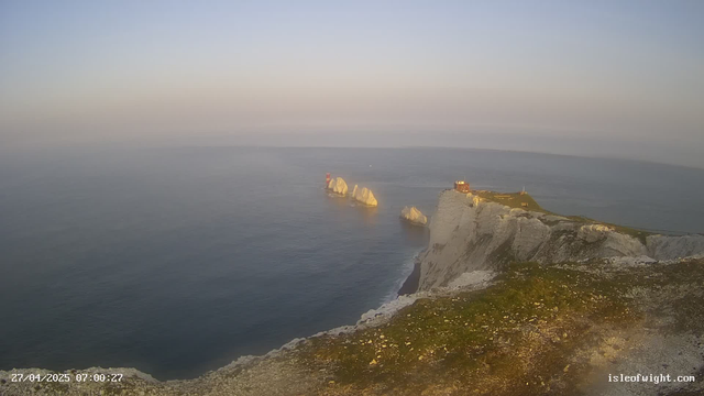 A view of white chalk cliffs and sea stacks along the coastline at sunrise, with a lighthouse on one of the cliffs and a building nearby. The sky is clear and the water is calm. The image is timestamped "27/04/2025 07:00:27" and has the website "isleofwight.com" in the corner.