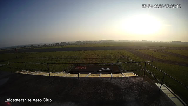 A wide view of a grassy airfield at sunrise, with a few small runways extending into the distance. The image appears to be taken from a webcam at Leicesterhire Aero Club, with the date and time displayed in the top right corner as April 27, 2025, at 07:00:45. The sky is clear with a bright sun near the horizon, casting a warm glow over the landscape. There are no aircraft or people visible in the image.