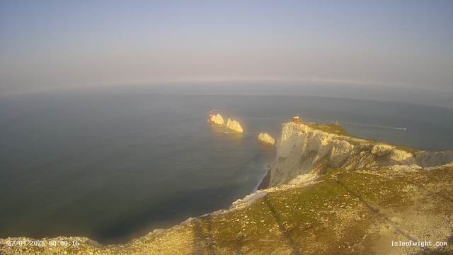A view from a webcam showing white chalk cliffs extending into the ocean, with a grassy area and a small building or lighthouse on top. Several large rocks are visible in the water near the cliffs. The scene appears to be during late afternoon or evening, with soft sunlight illuminating the cliffs and sea. The sky is clear with a gradient from blue to a faint haze near the horizon. The timestamp indicates it was taken on April 27, 2025, at 8:00:16.