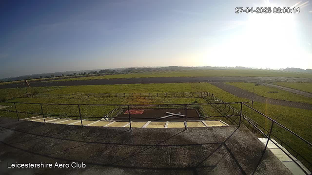 A view from a webcam showing an outdoor airfield with grassy areas, a paved taxiway, and a helipad with a large white "H" marked on it. The sky is clear with the sun shining brightly in the upper right corner. The scene is bathed in sunlight, creating a bright and open atmosphere. In the bottom left corner, there is text that reads "Leicestershire Aero Club," and in the upper right corner, there is a timestamp displaying the date and time: "27-04-2025 08:00:14."