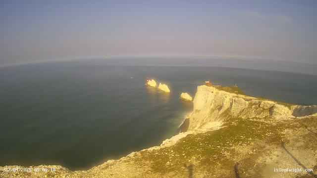 A panoramic view of a rugged coastline with tall white chalk cliffs extending into the ocean. There are several rock formations jutting out of the water, with a small lighthouse or navigation tower on one of the distant rocks. The sky is mostly clear with some thin clouds, and the water appears calm. The image is taken from an elevated vantage point on the cliffs, with a timestamp in the bottom left corner and the text "isledlight.com" in the bottom right corner.