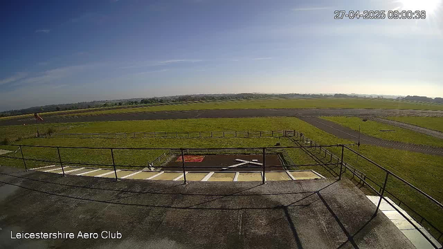 A wide view of an airfield taken from a webcam, showing a runway and taxiways in the distance, with grassy areas surrounding them. The sky is mostly clear with a few clouds, and the sunlight casts shadows on the ground. The timestamp in the top right corner indicates the date and time as April 27, 2025, at 9:00 AM. In the bottom left corner, the text reads "Leicestershire Aero Club."