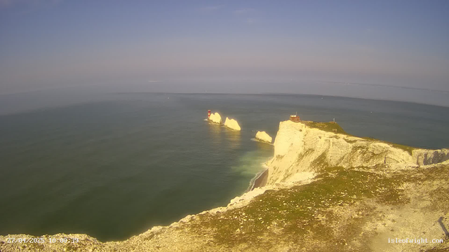 A panoramic view of white chalk cliffs along the coastline with a lighthouse on a smaller rocky island and a building on the main cliff. The sea below is calm, and the sky is clear with a light gradient from blue to a faint pinkish hue near the horizon. The scene is bathed in natural daylight.