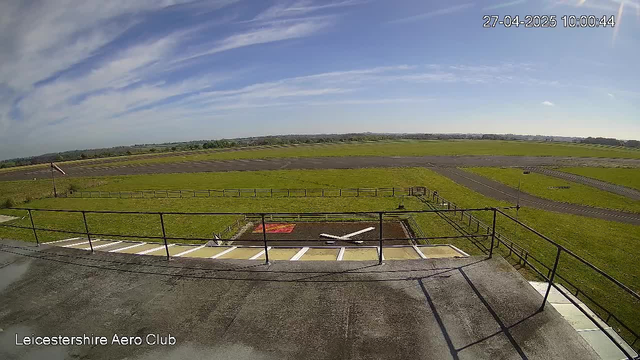 A wide view of an airfield with a clear blue sky and some thin, wispy clouds. In the foreground, there are parking spaces and a small fenced area with a windsock. The landscape extends into green fields and runways stretching into the distance. The date and time displayed in the upper right corner are "27-04-2025 10:00:44." The bottom left corner shows the text "Leicestershire Aero Club."