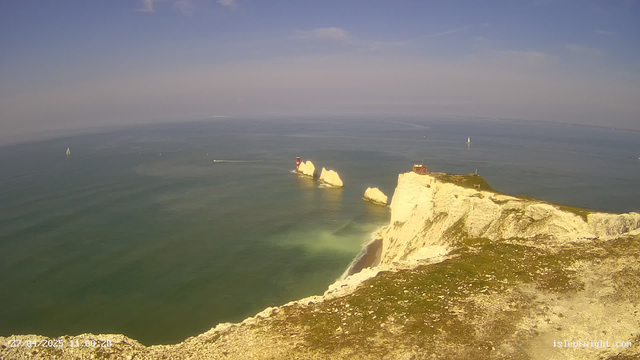 A scenic view of white chalk cliffs overlooking the ocean, with a small lighthouse and a building on top of the cliffs. Several sailboats are visible on the water, and the sky is mostly clear with some light clouds. The photo appears to be taken during the daytime.