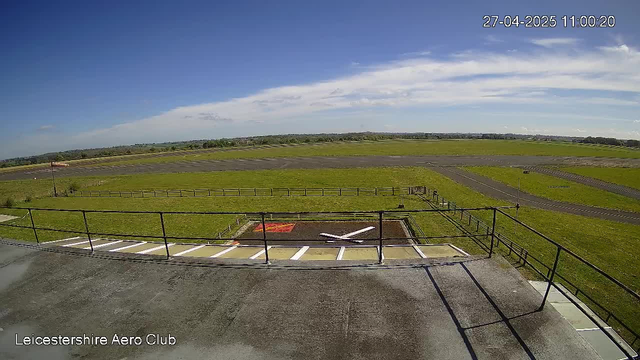 A view from a webcam at Leicestershire Aero Club showing an open grassy airfield with a taxiway and runway stretching into the distance under a clear blue sky with some scattered clouds. The foreground shows a helipad with painted markings and a small landing area, bordered by a metal railing. The top right corner displays a timestamp of "27-04-2025 11:00:20".
