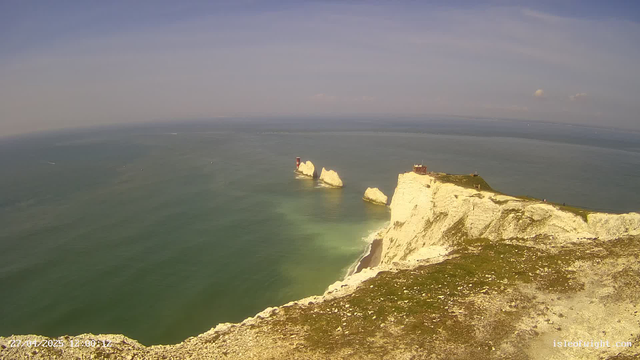 A view of tall white chalk cliffs overlooking the ocean, with a small building and a lighthouse situated at the edge of the cliff. The sea below is calm with a few boats visible in the distance, and the sky above is partly cloudy. The image is timestamped at the bottom left corner.