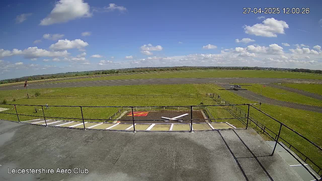 A view from a webcam at LeicesterShire Aero Club shows a bright, sunny day with a blue sky filled with fluffy white clouds. The foreground includes a balcony with a metal railing and painting lines on an adjacent concrete surface. Beyond the balcony, there is a green grassy area, a small helipad with a red border, and a paved taxiway leading to a runway. A small aircraft is taxiing on the runway, heading away from the viewer. The horizon line features a distant flat landscape with scattered trees. The date and time in the top right corner read "27-04-2025 12:00:26."