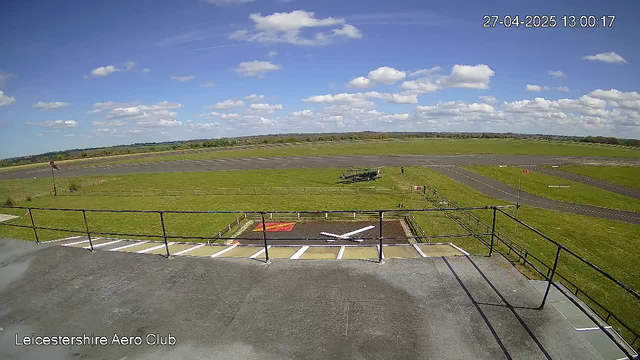 A wide view of an airfield on a bright, partly cloudy day. The foreground shows a fenced platform area with a small section marked with red and white lines. In the middle ground, there are open grassy fields along with a small airplane parked on the grass. The background features a long runway stretching into the distance, with a line of trees on the horizon. The sky is mostly blue with scattered fluffy white clouds. The top right corner displays the date and time as "27-04-2025 13:00:17". The bottom left corner indicates "Leicestershire Aero Club."