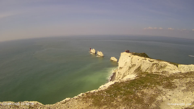 A wide view of white chalk cliffs extending into the ocean, with a lighthouse on one of the cliffs. The sea is calm with a few boats visible in the distance. The sky is mostly clear with some light clouds. The photo appears to be taken during the day with sunlight illuminating the landscape.