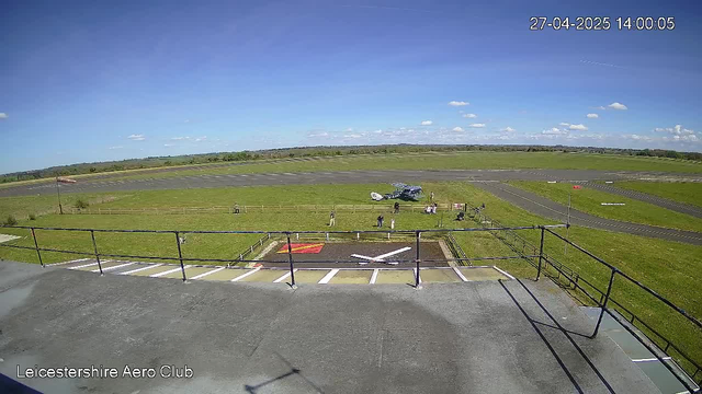 A view from a webcam at Leicestershire Aero Club shows a grassy airfield with a small airplane parked on the grass, several people walking or standing around, and a paved runway extending into the distance. The sky is clear with a few scattered clouds, and the scene is bathed in bright daylight. The date and time displayed in the top right corner are 27-04-2025, 14:00:05.