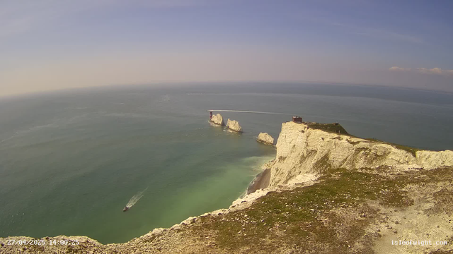 An aerial view of white chalk cliffs rising from the ocean, with a small building or structure on top of the cliffs. There are several large rock formations in the water near the base of the cliffs. A boat is sailing in the water, leaving a wake behind it. The sky is clear with a few clouds, and the overall scene is bright and sunny. The timestamp on the image indicates the date and time as April 27, 2025, at 14:00:25.