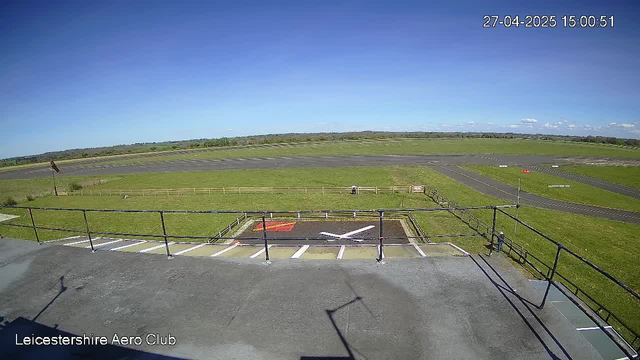 A view from a surveillance camera at Leicestershire Aero Club showing a clear blue sky, a grassy field, a distant runway, and a small helipad marked with a white "H" and red border in the foreground. The timestamp in the top right corner indicates the date and time as April 27, 2025, at 15:00:51.