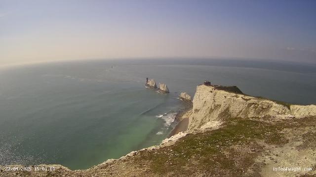 A panoramic view of white chalk cliffs overlooking the sea, with a grassy area on top. In the distance, there are large rock formations emerging from the water, including one with a small structure or tower on it. The sky is mostly clear with a slight haze near the horizon, and the water is a calm, muted greenish-blue. A sailboat or similar vessel is visible far out on the water. The date and time stamp shows "27/04/2025 15:01:01" in the bottom left corner.
