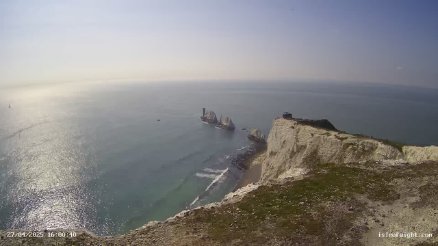 A wide view of white chalk cliffs overlooking the ocean, with a grassy top and a small building at the edge. In the distance, there are sea stacks and a lighthouse. The water reflects sunlight, creating a sparkling effect, and a few sailboats and small boats are visible on the water. The sky is mostly clear with a gentle haze near the horizon. The timestamp shows "27/04/2025 16:00:40" and the website "isleduight.com" is displayed in the bottom right corner.