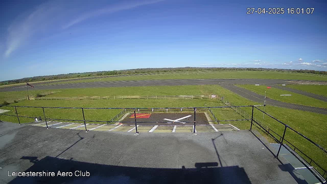 A view from a webcam at Leicesterhire Aero Club showing a clear blue sky, an empty tarmac runway with some distant runways, green grass fields, and a windsock on the left. The date and time are displayed in the top right corner, and the club's name is at the bottom left.
