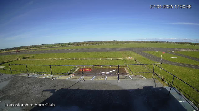 An empty parking area with designated slots, viewed from a higher vantage point, with a green field and runway extending into the distance under a clear blue sky. The image is timestamped "27-04-2025 17:00:06" and labeled "Leicestershire Aero Club" at the bottom left corner.