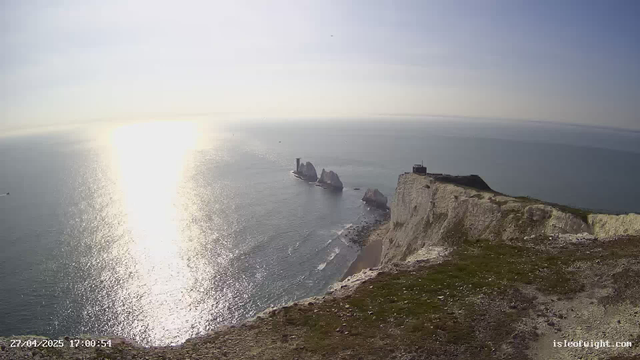 A scenic view of white chalk cliffs overlooking the ocean during sunset, with the sunlight reflecting on the water. A lighthouse and a small building are visible on top of the cliff to the right, and several rock formations extend into the sea in the distance. The foreground shows a grassy and rocky edge of the cliff. The timestamp at the bottom left indicates the date and time as April 27, 2025, at 5:00:54 PM.