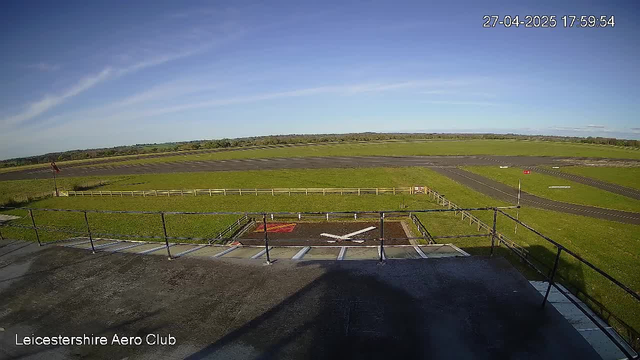 A wide-angle view of a small airfield with a runway stretching into the distance under a clear blue sky. In the foreground, there is a helipad with a white helicopter symbol and a red sign. The surrounding grassy area is lush and green, bordered by a wooden fence. There are no aircraft or people visible in the image. The timestamp in the upper right corner reads "27-04-2025 17:59:54," and the bottom left corner has the text "Leicestershire Aero Club."