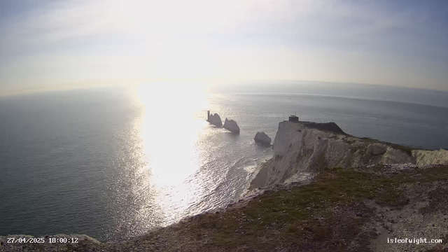 A view of white chalk cliffs along the coastline at sunset, with the sun reflecting off the calm sea. There is a small building or structure on top of the cliffs, and several large rocks or sea stacks jutting out into the water. The date and time are displayed at the bottom left corner, and a website URL is in the bottom right corner.