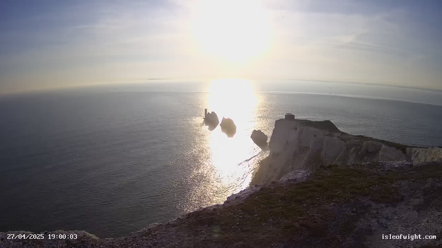 A view of a coastal cliff at sunset with the sun low on the horizon, reflecting bright light on the calm ocean waters below. There are notable rock formations and structures on the edge of the cliff, and the sky is partly cloudy with a peaceful, scenic atmosphere. The timestamp in the bottom left corner reads "27/04/2025 19:00:03," and the website "isleofwight.com" is displayed in the bottom right corner.