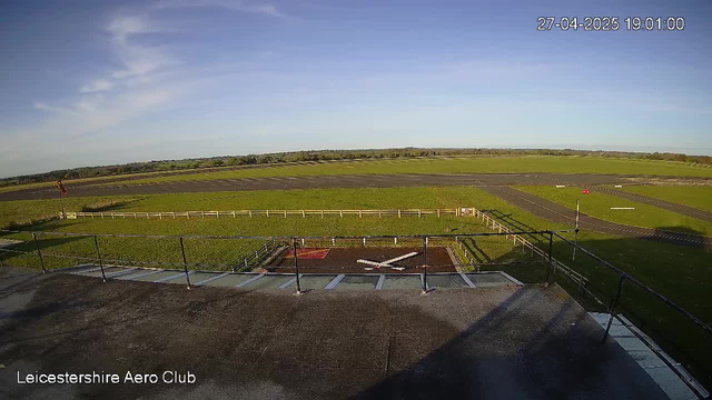 An empty airfield runway with a large open grassy area beyond, bordered by a low white fence. The sky is mostly clear with some wispy clouds, and the sun is shining brightly. The date and time are displayed in the top right corner as "27-04-2025 19:01:00", and the bottom left corner has the text "Leicestershire Aero Club."