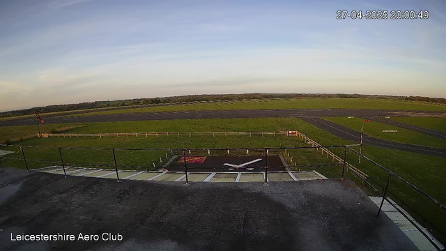 An outdoor view from a webcam at Leicesterhire Aero Club showing a clear sky with a few wispy clouds, a spacious grassy area encircled by a white fence, and a paved runway winding through the landscape. The timestamp reads 27-04-2025 20:00:49.
