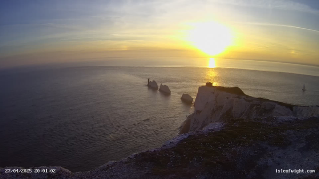 A sunset over the ocean with white chalk cliffs in the foreground. The sun is low on the horizon, casting a bright reflection on the water. There are a few boats visible in the distance, and one prominent structure on top of a cliff. The scene has a peaceful and scenic atmosphere.