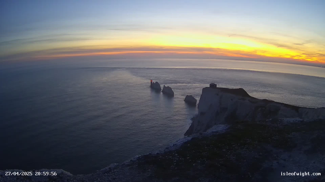 A view of the ocean at sunset, featuring white chalk cliffs with dark rocks and a small house or structure on top. The sky displays a gradient of colors from yellow to orange and blue, with some clouds near the horizon. There are three prominent sea stacks with a lighthouse on one of them, and a trail of water from a boat or ship moving through the water. The timestamp in the bottom left corner is "27/04/2025 20:59:56," and a website "isleofwight.com" is visible in the bottom right corner.