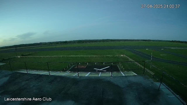 An outdoor view from a webcam at Leicesterhire Aero Club shows a section of a runway and surrounding grassy fields under a cloudy sky. The timestamp reads 27-04-2025 21:00:37 in the top right corner.