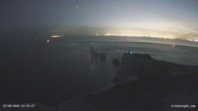 A dark, nighttime scene of a coastal area with a faint horizon line and a distant city or village visible on the horizon. The sky is partly cloudy with some stars visible, and there are a few small lights in the water, possibly from boats or buoys. The shoreline is visible in the foreground, with a rocky or uneven surface. A timestamp in the lower left corner reads "27/04/2025 21:59:27," and a website URL "isleofwight.com" is displayed in the lower right corner.