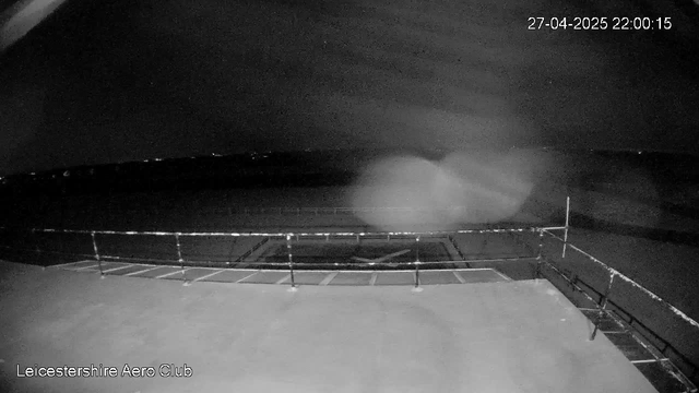 A black-and-white image from a webcam shows an empty outdoor area with a railing or fence in the foreground. The background reveals a dark, open space with a faint, cloudy outline of a large object or cloud formation in the sky. The timestamp in the upper right corner indicates the date and time as 27th April 2025 at 22:00:15. The words "Leicestershire Aero Club" are displayed in the lower left corner.