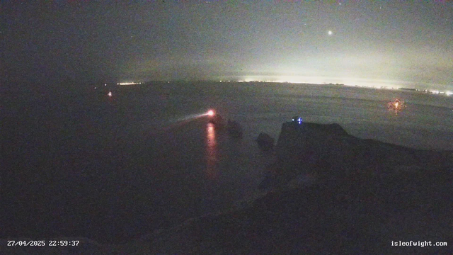 A dark scene at night with faint reflections on the water surface. Several small lights are visible on the horizon and on boats or structures in the water, with some reflections scattered across the surface. The sky shows a faint bright spot, possibly the moon or bright star. The timestamp in the bottom left corner reads "27/04/2025 22:59:37", and there is a website reference "isleofwight.com" in the bottom right corner.