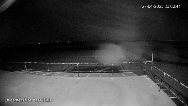 A black-and-white image from a webcam shows a dark, open outdoor area with a railing or barrier visible in the foreground. The background features a mostly clear sky with some faint lights on the horizon. A timestamp in the top right corner indicates the date as April 27, 2025, and the time as 23:00:41. The bottom left corner displays the text "Leicestershire Aero Club."