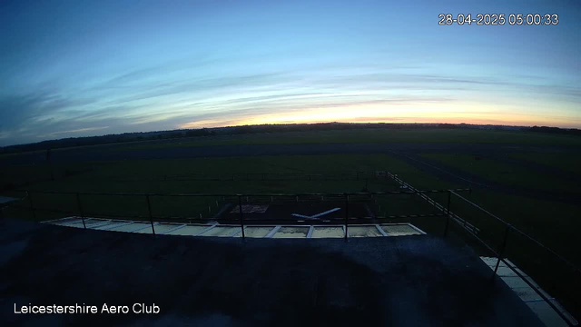 A wide view of an airfield at dawn, with a clear sky and a subtle gradient of colors from dark blue to light near the horizon. In the foreground, there is a platform with railings and a small aircraft on a taxiway. The image includes a timestamp in the top right corner reading "28-04-2025 05:00:33" and the text "Leicestershire Aero Club" at the bottom left.