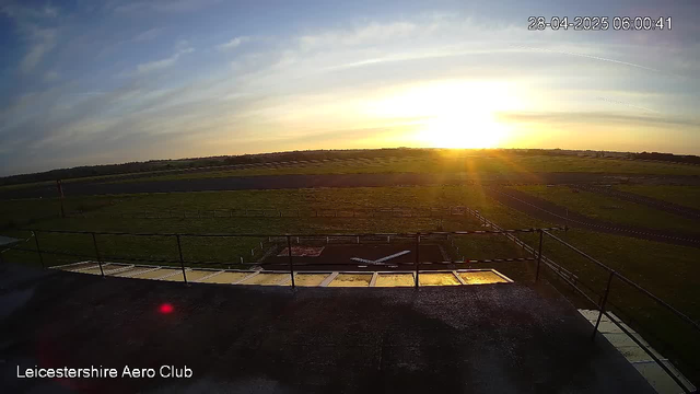 A sunrise or sunset over an open, grassy airfield with a clear sky and some wispy clouds. The image shows a flat landscape with a runway stretching into the distance. There are safety barriers and some structures on a platform in the foreground. The timestamp in the top right indicates the date and time as April 28, 2025, at 6:00:41 AM. The bottom left corner has the text "Leicestershire Aero Club."
