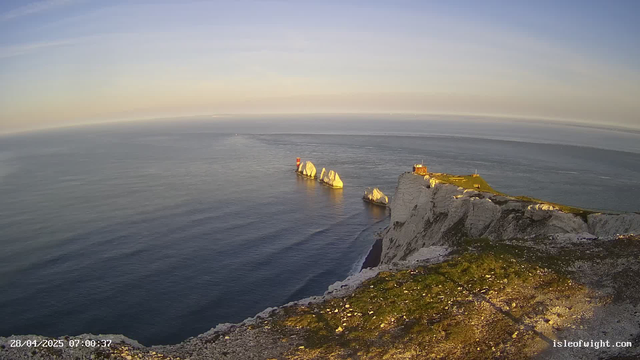 An aerial view of white chalk cliffs overlooking the ocean at sunrise, with patches of green grass on top and a small building near the edge. Several large white rocks are visible in the water near the base of the cliffs. The sky is mostly clear with subtle pastel hues. The website URL "isleofwight.com" is displayed in the bottom right corner.