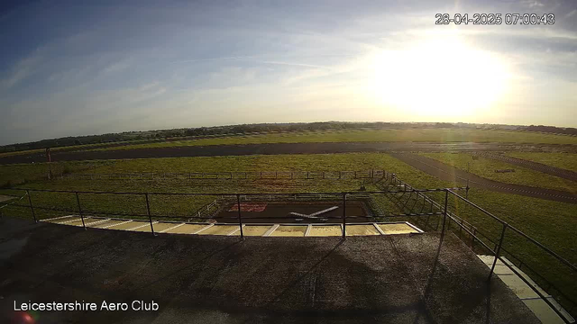An early morning view from a webcam at Leicestershire Aero Club shows a grassy airfield with a small aircraft parked on the tarmac. The sky is clear with a few wispy clouds, and the sun is shining brightly near the horizon, creating a warm glow. The image is timestamped "28-04-2025 07:00:43" in the top right corner. A railing surrounds the platform from which the photo is taken, and the landscape extends to rolling fields and distant horizon. The words "Leicestershire Aero Club" are displayed at the bottom left corner.
