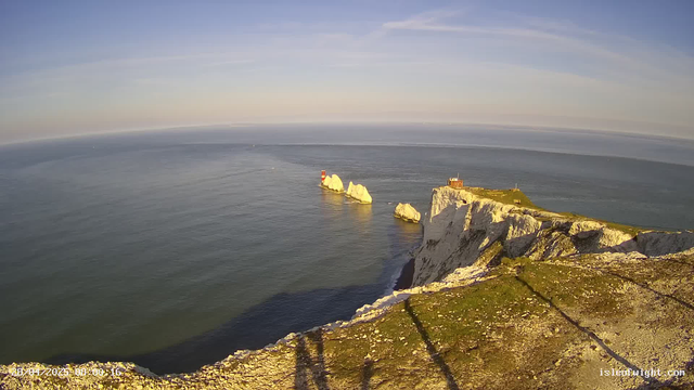 A panoramic view of white chalk cliffs on the coast with a grassy top, extending into the sea. Several large white rocks are visible in the water near the cliffs. A small building or lighthouse is situated on the grassy area of the cliffs. The water is calm, reflecting the sky, which is mostly clear with some light clouds, and the scene appears to be taken during a sunny day, possibly in the late afternoon or early evening based on the warm lighting. Shadows from a railing or barrier are cast onto the ground in the foreground.