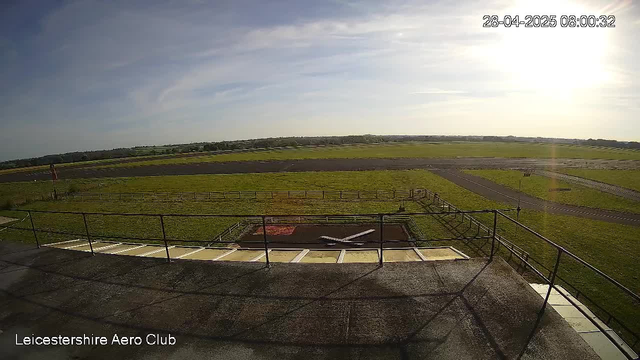 A wide view of an open grassy airfield with taxiways and runways stretching into the distance. The sky is mostly clear with some light clouds, and the sun is shining brightly from the upper right corner. On the bottom left, there is text that reads "Leicestershire Aero Club." The image is timestamped in the top right corner with "28-04-2025 08:00:32."
