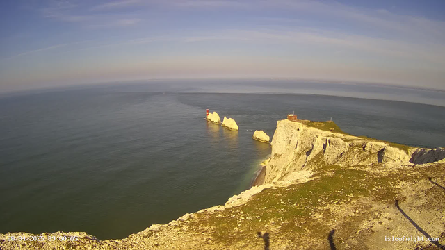 A scenic view of white chalk cliffs with a grassy top, overlooking the ocean. Several large white rocks are visible in the water near the base of the cliffs. A red and white lighthouse is positioned on one of the rocks in the distance. The sky is mostly clear with some wispy clouds. The time and date are displayed at the bottom left corner: 28/04/2023 at 09:00:05.