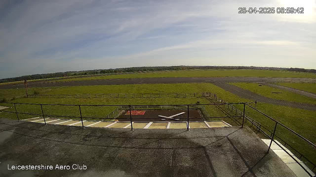 A view from a webcam showing an airfield with a runway in the distance, surrounded by green grass and open sky with some wispy clouds. The timestamp on the upper right corner indicates it was taken on April 28, 2025, at 8:59 AM. The bottom left corner displays the text "Leicestershire Aero Club." There are railings and a ladder-like structure in the foreground, and the overall scene suggests a quiet, rural airport setting.