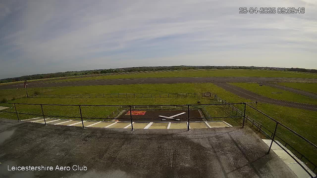 A wide view of an outdoor airfield shows a tarmac area with a small helicopter pad and a railing in the foreground. Beyond, there are grassy fields and multiple runways stretching into the distance under a partly cloudy sky. The timestamp indicates the date as April 28, 2025, at 9:59 AM. The bottom left corner has the text "Leicestershire Aero Club."