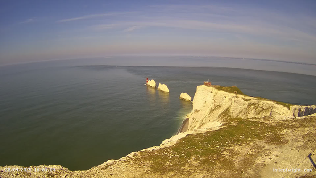 A view from a webcam shows white chalk cliffs along a coastline, with a grassy top. In the water below, there is a small lighthouse and several large white rock formations. The sky is mostly clear with a few wispy clouds, and the calm sea reflects the sky's light colors. The photo is timestamped "28/04/2023 10:00:04" in the bottom left corner.