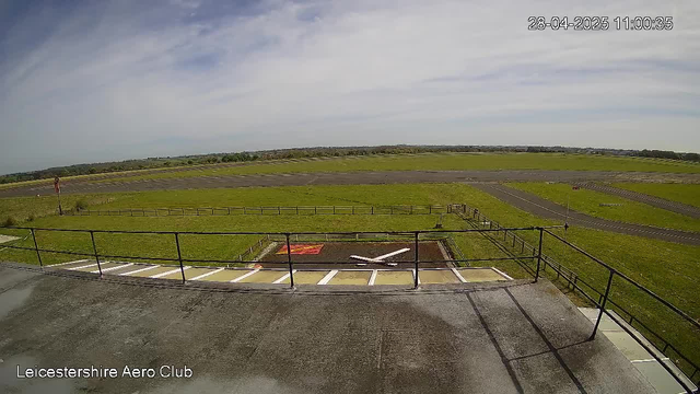 A wide view of an airfield with a grassy area, runways, and taxiways stretching into the distance under a partly cloudy sky. A small parking area with marked spaces and a ramp with safety railings are in the foreground. The timestamp in the top right corner shows "28-04-2025 11:00:35," and the bottom left corner displays "Leicestershire Aero Club."
